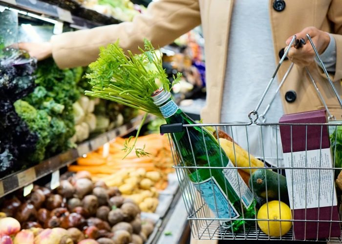 Woman selecting fresh produce in a supermarket with a basket full of groceries.